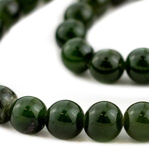 May include: A necklace of dark green, spherical beads, shown close-up. The beads have a polished, glossy surface, reflecting light. The necklace is set against a white backdrop, accentuating the colour and texture of the beads.