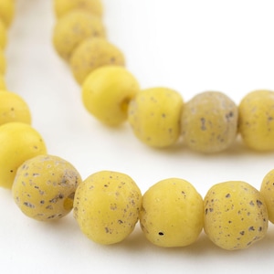 May include: Close-up of a strand of yellow beads with a speckled pattern. The beads are round and have a matte finish. The beads are strung together, creating a necklace or bracelet.