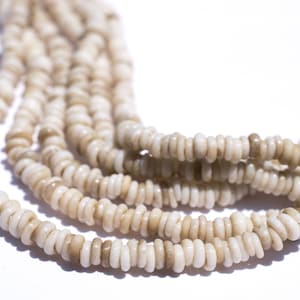 Könnte beinhalten: Nahaufnahme von mehreren Strängen aus scheibenförmigen, cremefarbenen Perlen. Die Perlen haben ein natürliches, organisches Aussehen mit Farbvariationen, die von hellem Creme bis zu blassem Braun reichen. Die Stränge sind in einem fließenden, geschwungenen Muster vor weißem Hintergrund angeordnet.