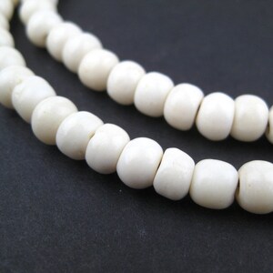 May include: Close-up of two strands of off-white, round beads. The beads are strung together, with a dark brown string visible at the end. The background is a solid, dark gray color.