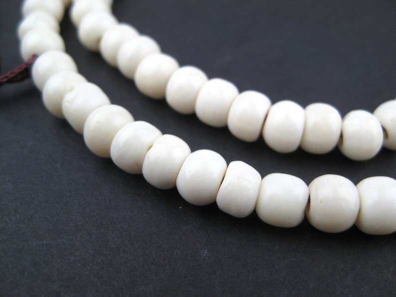 May include: Close-up of two strands of off-white, round beads. The beads are strung together, with a dark brown string visible at the end. The background is a solid, dark gray color.