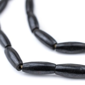 Puede incluir: Primer plano de un collar de cuentas de madera negras. Las cuentas son de forma oblonga y están ensartadas, creando un collar o una pulsera. El color oscuro de las cuentas contrasta con el fondo blanco, resaltando la veta natural de la madera.