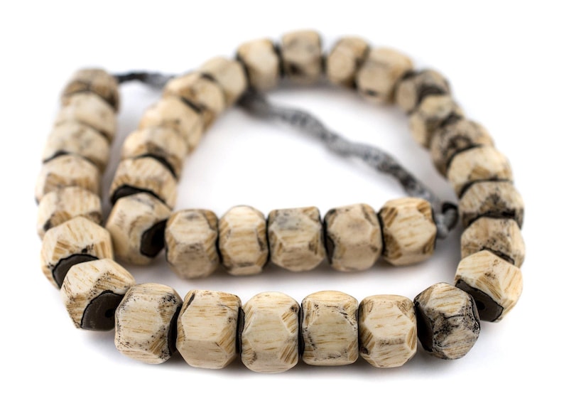 May include: A string of beige bone beads with a dark grey string threaded through them. The beads are faceted and have a worn, rustic look.