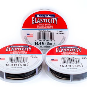 Puede incluir: Tres carretes de cord&oacute;n el&aacute;stico Beadalon Elasticity negro, cada uno con una etiqueta blanca y una pancarta roja que dice "Beadalon Elasticity". Las etiquetas indican el grosor del cord&oacute;n en pulgadas y mil&iacute;metros, y la longitud del cord&oacute;n en pies y metros. Los carretes est&aacute;n sobre una superficie blanca.