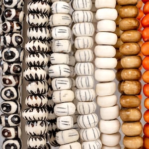 May include: Close-up of several strands of beads in various colors and patterns. The beads include black and white patterned, white with black stripes, light brown, and orange. The beads are round and strung together.