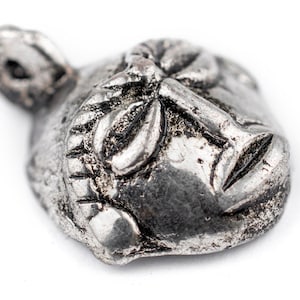 May include: A silver pendant in the shape of a face with detailed features. The face has a closed mouth and eyes, and a textured surface.