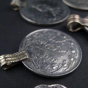 May include: A silver coin with Arabic script and a gold-colored loop on the side. The coin has a worn, aged appearance.