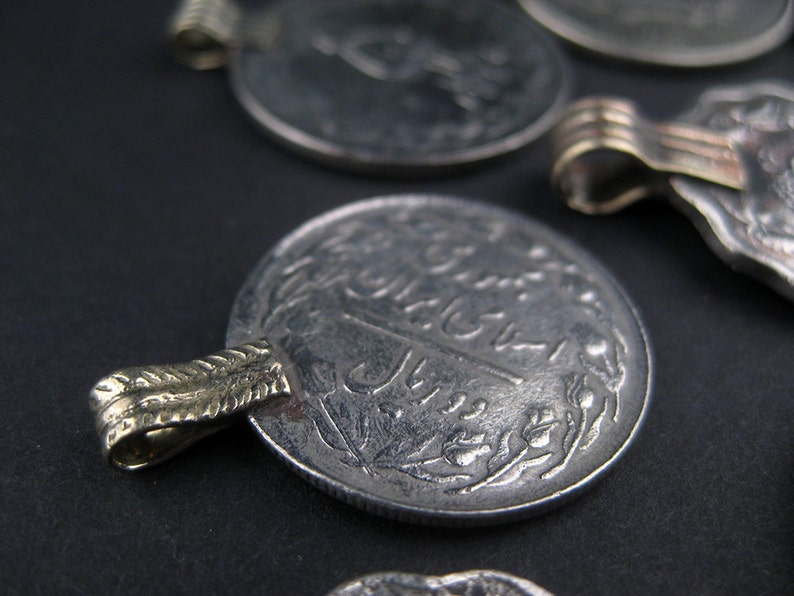 May include: A silver coin with Arabic script and a gold-colored loop on the side. The coin has a worn, aged appearance.