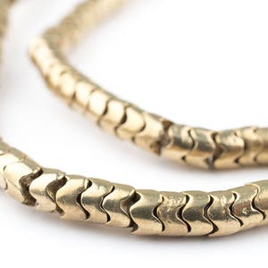 Może przedstawiać: Zbliżenie złotego naszyjnika o unikalnym, teksturowanym wzorze. Naszyjnik składa się z zazębiających się ogniw w kształcie szewronów, tworząc elastyczny i przyciągający wzrok element. Ciepły złoty odcień kontrastuje z białym tłem.