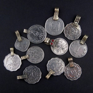 May include: A collection of 11 silver and gold pendants, each featuring a different design. The pendants are shaped like coins and have a variety of intricate patterns and symbols. Some of the pendants have red gemstones.