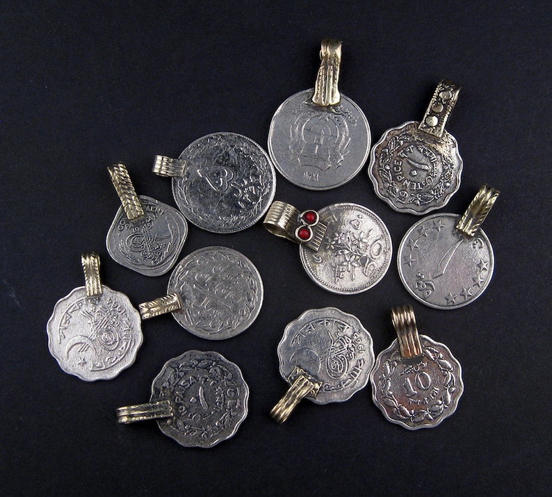 May include: A collection of 11 silver and gold pendants, each featuring a different design. The pendants are shaped like coins and have a variety of intricate patterns and symbols. Some of the pendants have red gemstones.