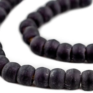 Puede incluir: Primer plano de un collar de cuentas moradas oscuras. Las cuentas son cilíndricas con una textura ligeramente rugosa y están ensartadas en un cordón fino y oscuro. El fondo es blanco liso.