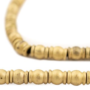 May include: Close-up of a gold-colored beaded necklace. The necklace features a series of small, round beads and larger, cylindrical beads. The beads are strung together on a dark cord, creating a simple yet elegant design.