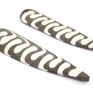 May include: Two elongated, dark gray pendants with a white, wavy pattern. Each pendant has a small hole at the top for stringing. The pendants are displayed against a white background, highlighting their unique design.