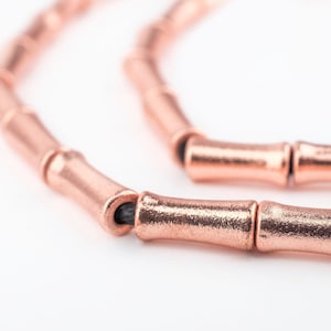 Pode incluir: Um close-up de um bracelete de contas de estilo bambu em ouro rosa. As contas são conectadas por um cordão elástico preto.