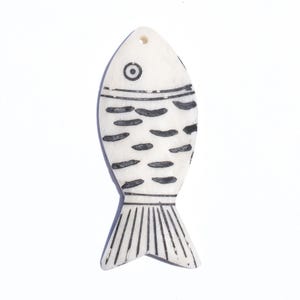 May include: A white, fish-shaped pendant with black detailing. The pendant features a simple eye, horizontal lines, and curved shapes to represent scales. The tail has vertical lines. A small hole is at the top for stringing.