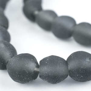 May include: A string of matte grey beads. The beads are oval-shaped and have a slightly textured surface. The beads are strung together, creating a necklace or bracelet. The background is white.