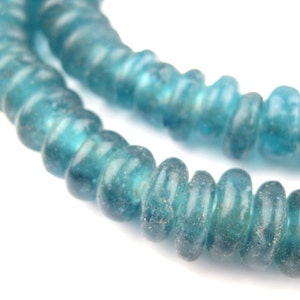 May include: Close-up of a strand of turquoise-colored, irregular glass beads. The beads have a slightly rough texture and are strung together, creating a curved line against a white background. The beads are a translucent blue-green color.