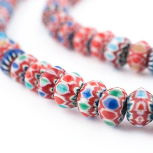 Puede incluir: Primer plano de un collar de cuentas de vidrio de colores. Las cuentas son principalmente rojas, blancas, azules y verdes, con patrones intrincados. Las cuentas están ensartadas, creando una línea curva sobre un fondo blanco.