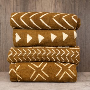 May include: Four folded brown fabric textiles with white geometric patterns. The patterns include chevrons, triangles, and cross-like designs. The textiles are stacked on top of each other, showcasing the different patterns.