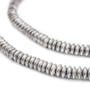 K&ouml;nnte beinhalten: Nahaufnahme von zwei Str&auml;ngen silberfarbener, scheibenf&ouml;rmiger Perlen. Die Perlen sind aufgef&auml;delt und bilden eine Halskette oder ein Armband. Die Perlen haben eine strukturierte, matte Oberfl&auml;che vor wei&szlig;em Hintergrund.