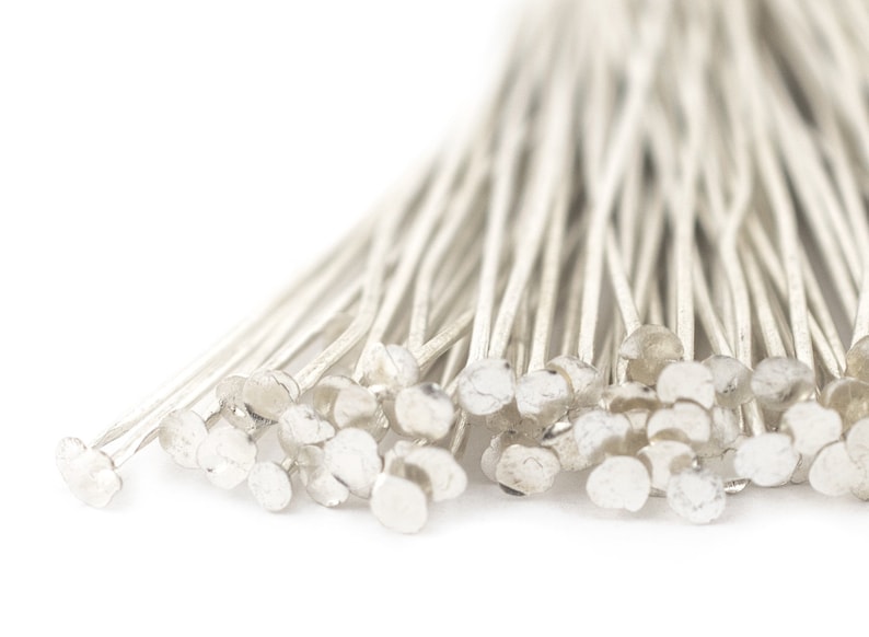 May include: A close-up of a bunch of silver head pins. The pins are arranged in a fan shape, with the heads of the pins facing the viewer. The pins are all the same size and shape, and they have a smooth, shiny finish.