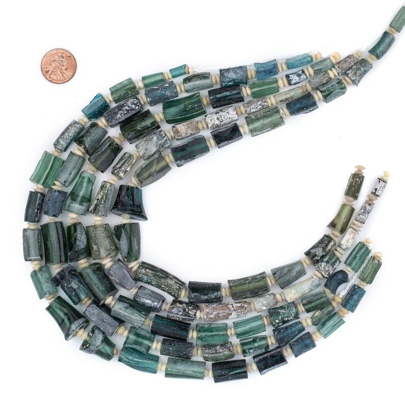 May include: A strand of green and blue glass beads with white spacers. The beads are cylindrical and have a slightly irregular shape. The beads are strung on a cord and are arranged in a loose, flowing pattern.