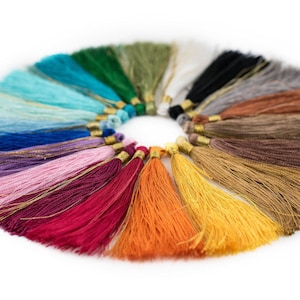 May include: A collection of 20 colourful tassels arranged in a circle. The tassels are made of soft yarn and have a gold-coloured metal top. The colours include teal, blue, purple, pink, red, orange, yellow, brown, grey, and white.