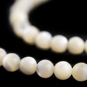 May include: Close-up of a strand of off-white, round beads against a black background. The beads are smooth and evenly sized, suggesting a necklace or bracelet. The lighting highlights the subtle luster of the beads.