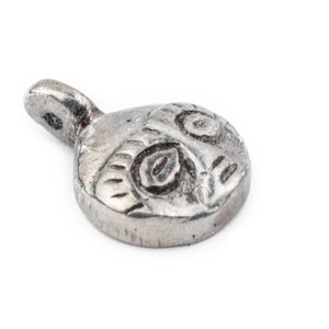 May include: A small, silver-toned pendant with a stylized owl design. The pendant features a circular body with engraved details depicting the owl's face, including eyes and feathers. A small loop extends from the top for attachment.