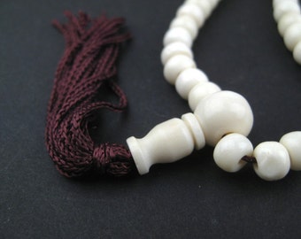 May include: Ivory-colored prayer beads with a dark burgundy tassel. The beads are round and smooth, with a larger bead at the center. The tassel is made of tightly woven threads. The background is a solid black.