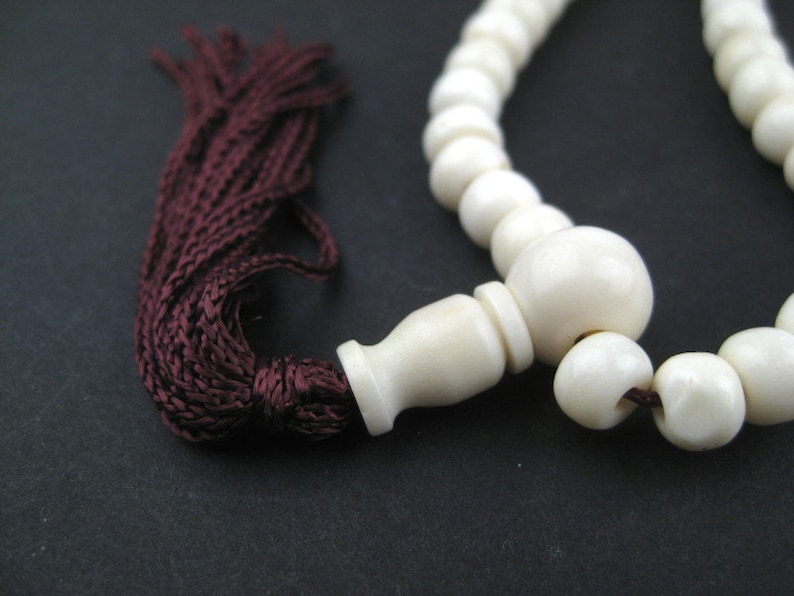 May include: Ivory-colored prayer beads with a dark burgundy tassel. The beads are round and smooth, with a larger bead at the center. The tassel is made of tightly woven threads. The background is a solid black.