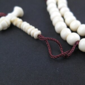 May include: Ivory-colored prayer beads with a tassel and a decorative end piece. The beads are strung on a dark red cord. The background is a solid black.