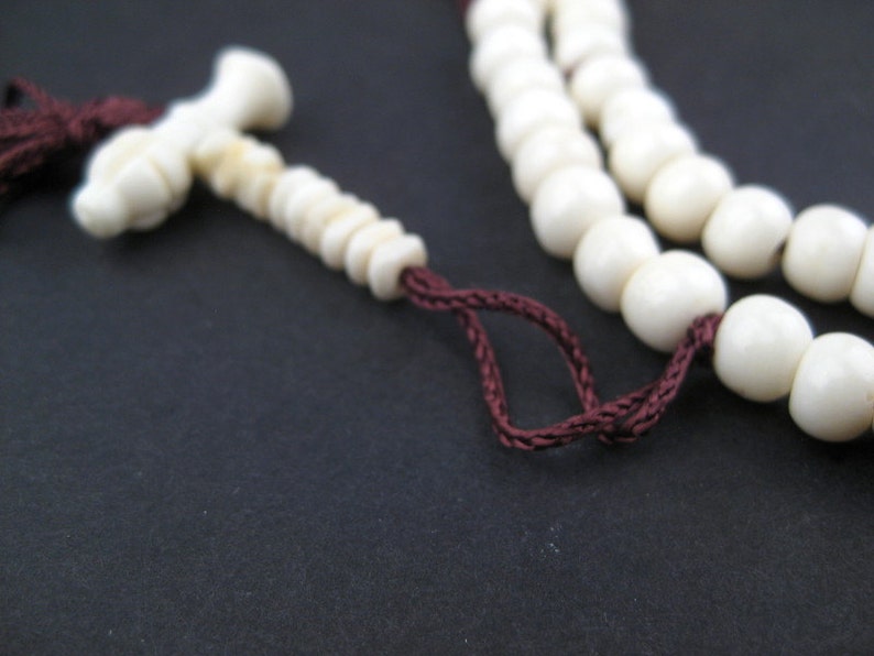 May include: Ivory-colored prayer beads with a tassel and a decorative end piece. The beads are strung on a dark red cord. The background is a solid black.