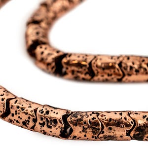 May include: Close-up of a copper-colored necklace with a textured, arrow-shaped design. The necklace is made of individual, connected segments, creating a flexible chain. The metal has a distressed finish, adding to its unique appeal.