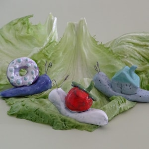 Könnte beinhalten: Drei Keramik-Schnecken mit verschiedenen Dekorationen. Die erste Schnecke ist blau mit einem weißen und türkisfarbenen Donut auf dem Rücken. Die zweite Schnecke ist weiß mit einer roten Erdbeere auf dem Rücken. Die dritte Schnecke ist blau mit einem hellblauen Haus auf dem Rücken. Die Schnecken befinden sich auf einem grünen Salatblatt.