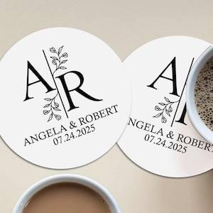 Può includere: Due sottobicchieri rotondi bianchi con disegni floreali neri e le iniziali "A" e "R" intrecciate. Il testo "Angela & Robert 07.24.2025" è stampato su ogni sottobicchiere.