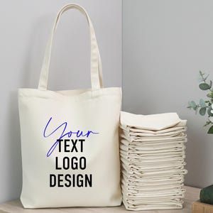以下が含まれることがあります： 長いハンドルが付いたクリーム色のトートバッグで、青と黒で「Your Text Logo Design」というテキストが特徴です。折りたたまれたクリーム色の生地の山がバッグの隣にあります。背景には白い花の入った花瓶があります。
