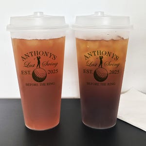 May include: Two clear plastic cups with black lids filled with a dark brown liquid. The cups have a black and gold design with the text "Anthony's Last Swing Est. 2025 Before the Ring".