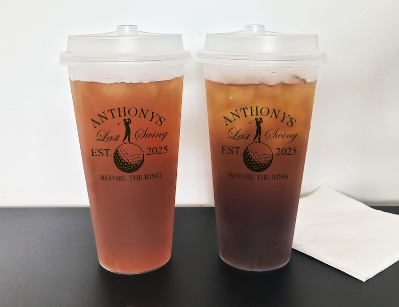 May include: Two clear plastic cups with black lids filled with a dark brown liquid. The cups have a black and gold design with the text "Anthony's Last Swing Est. 2025 Before the Ring".