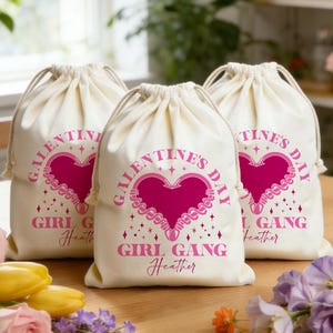 May include: Three off-white drawstring bags with pink text and heart designs. The bags read "GALENTINE'S DAY GIRL GANG" with a name below. The bags are on a wooden surface with flowers.