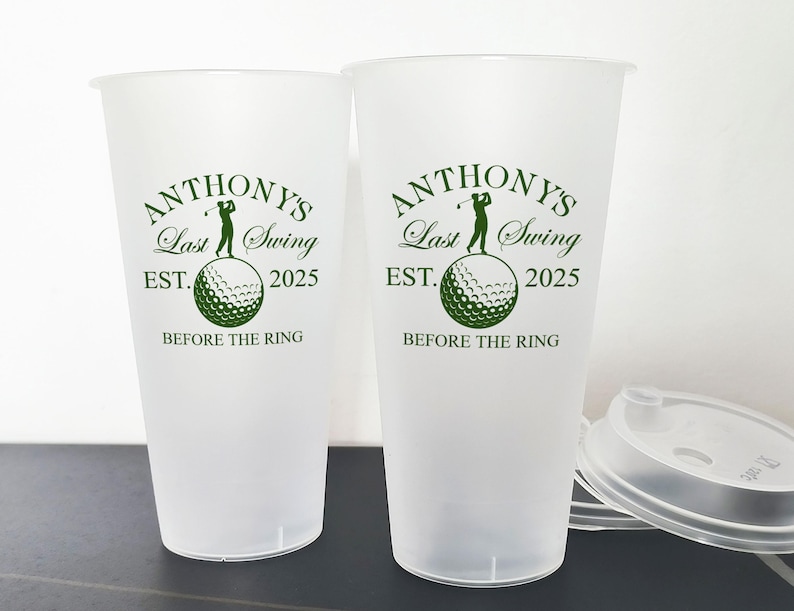 May include: Two white plastic cups with green text and a golf ball graphic. The text reads "Anthony's Last Swing Est. 2025 Before the Ring".