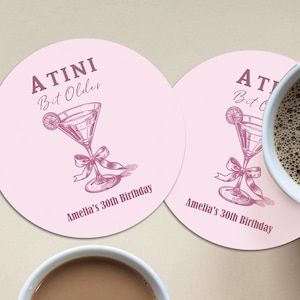 May include: Two pink round coasters with a martini glass design and the text "A TINI Bit Older" and "Amelia's 30th Birthday".