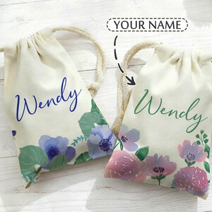 May include: Two off-white drawstring bags with the name "Wendy" printed in blue and green script. The bags are decorated with watercolor floral designs in shades of purple, blue, and pink, with green leaves. The bags have beige rope drawstrings.