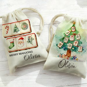 May include: Two off-white drawstring bags with Christmas-themed watercolour designs. One bag features Mahjong tiles with festive images, the text "MERRY MAHJONG" and the name "Olivia". The other bag has a Christmas tree made of Mahjong tiles and the name "Olivia".