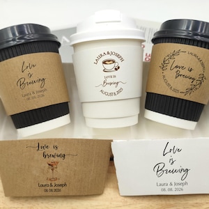 Puede incluir: Tres tazas de café con tapas negras y mangas marrones, y una taza blanca con tapa blanca. Las mangas y las tazas están impresas con las palabras "Love is Brewing" y nombres y fechas. Las tazas están sobre una bandeja blanca.
