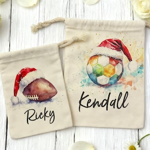 Op de afbeelding: Twee beige trekkoordzakken met aquarel illustraties. Eén toont een American football met een kerstmuts en de naam "Ricky". De andere zak toont een voetbal met een kerstmuts en de naam "Kendall".