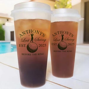 May include: Two clear plastic cups with lids filled with a brown liquid. The cups have a black and white graphic with the text "Anthony's Last Swing Est. 2025 Before the Ring".
