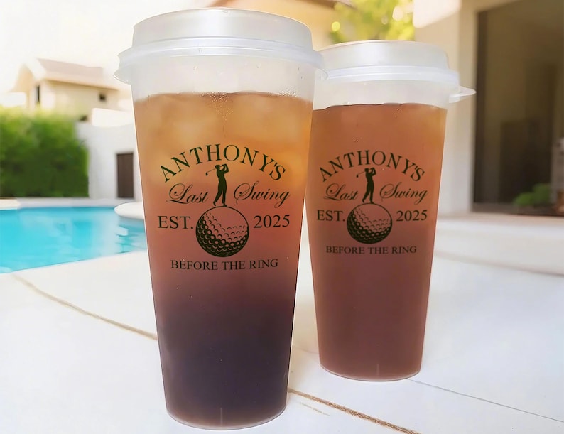 May include: Two clear plastic cups with lids filled with a brown liquid. The cups have a black and white graphic with the text "Anthony's Last Swing Est. 2025 Before the Ring".