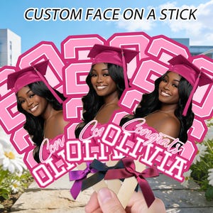Puede incluir: Recortes de caras personalizados en palos con una mujer sonriente con birrete. Los recortes son rosas con el año "2023" y el nombre "Olivia" en blanco. El texto "Custom Face on a Stick" está en la parte superior.